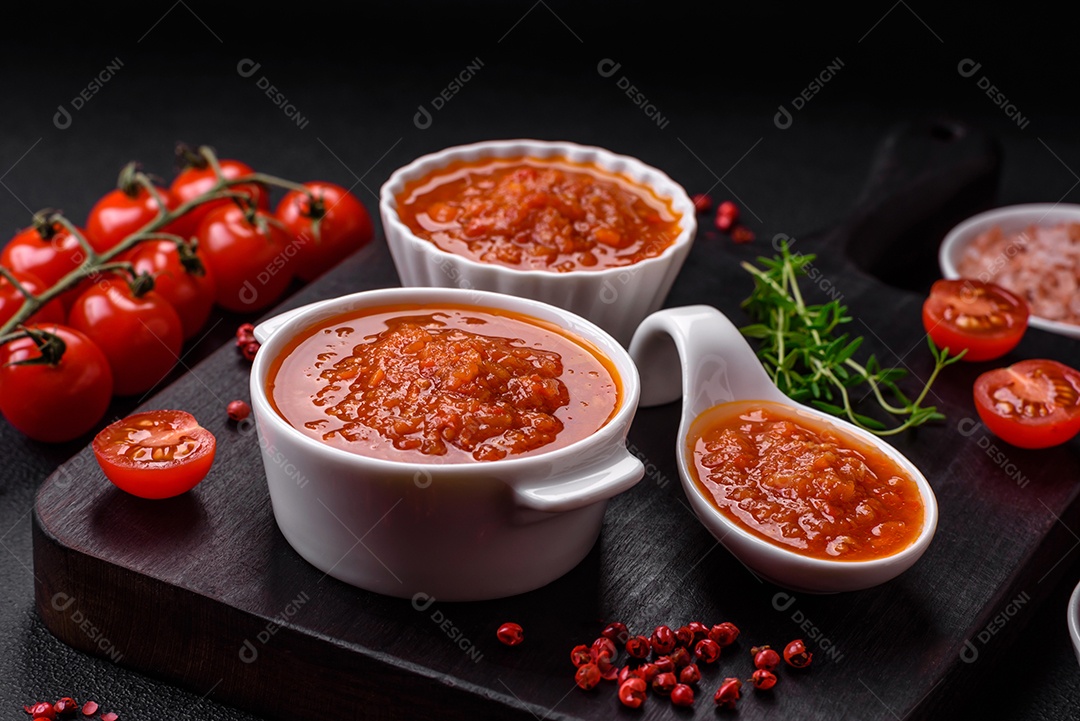 Delicioso molho de tomate picante com pimenta, alho, sal, especiarias e ervas em uma molheira branca sobre um fundo escuro de concreto