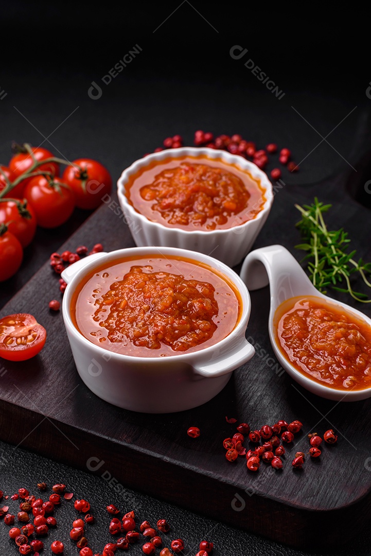 Delicioso molho de tomate picante com pimenta, alho, sal, especiarias e ervas em uma molheira branca sobre um fundo escuro de concreto