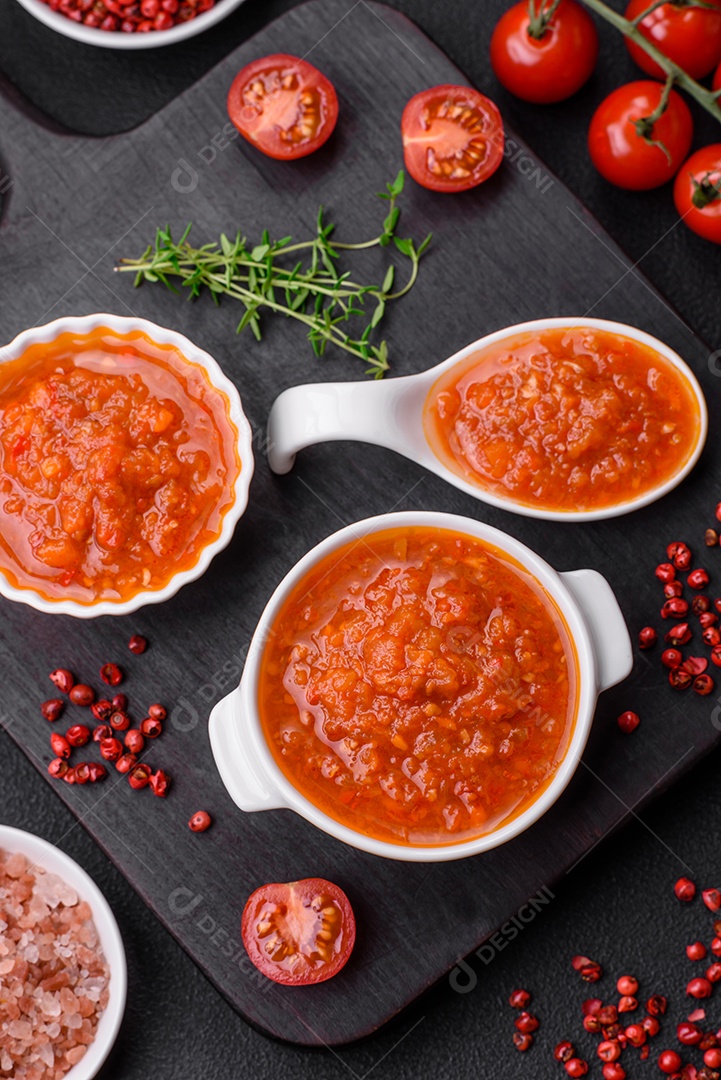 Delicioso molho de tomate picante com pimenta, alho, sal, especiarias e ervas em uma molheira branca sobre um fundo escuro de concreto