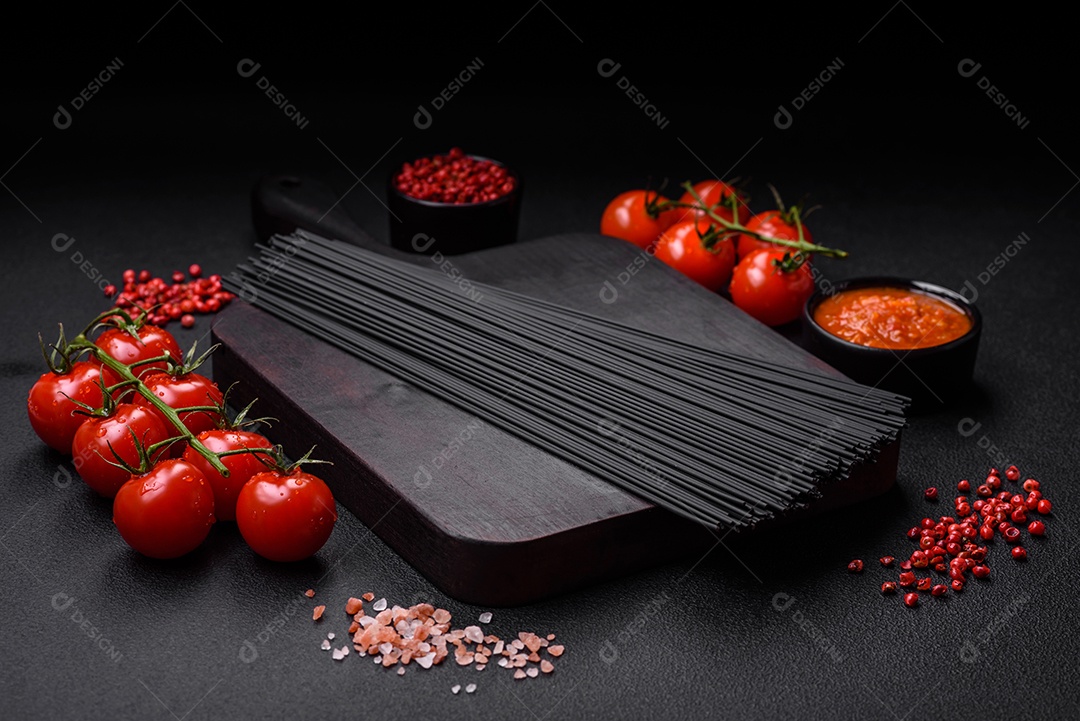 Massa de espaguete preta crua com tomate, sal, especiarias e ervas sobre um fundo escuro de concreto