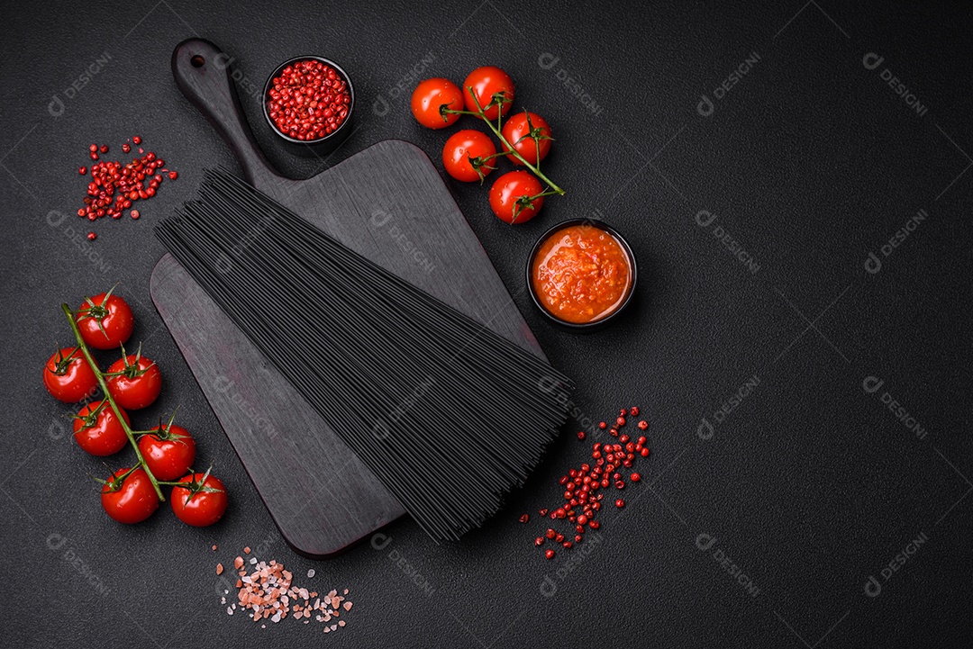 Massa de espaguete preta crua com tomate, sal, especiarias e ervas sobre um fundo escuro de concreto