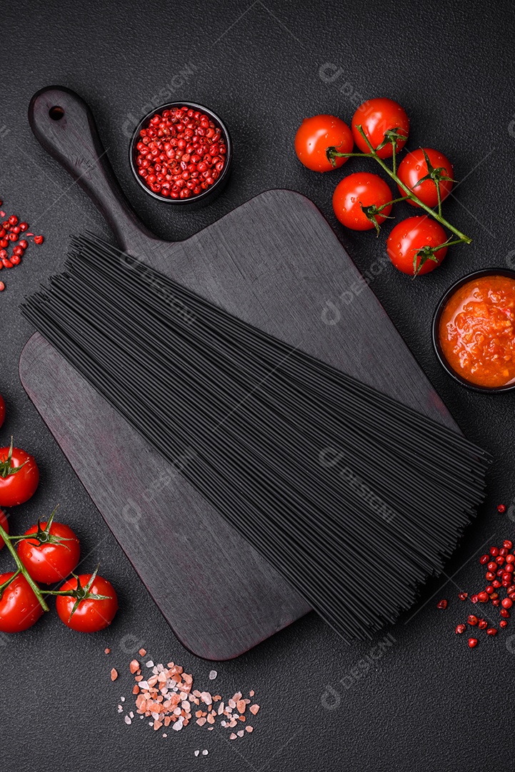 Massa de espaguete preta crua com tomate, sal, especiarias e ervas sobre um fundo escuro de concreto