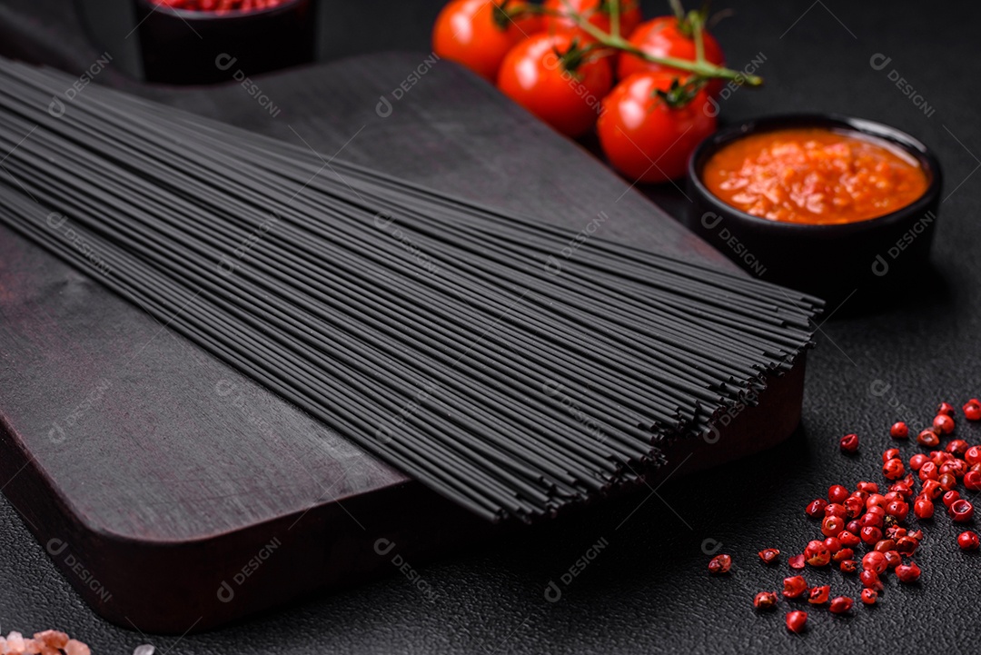 Massa de espaguete preta crua com tomate, sal, especiarias e ervas sobre um fundo escuro de concreto