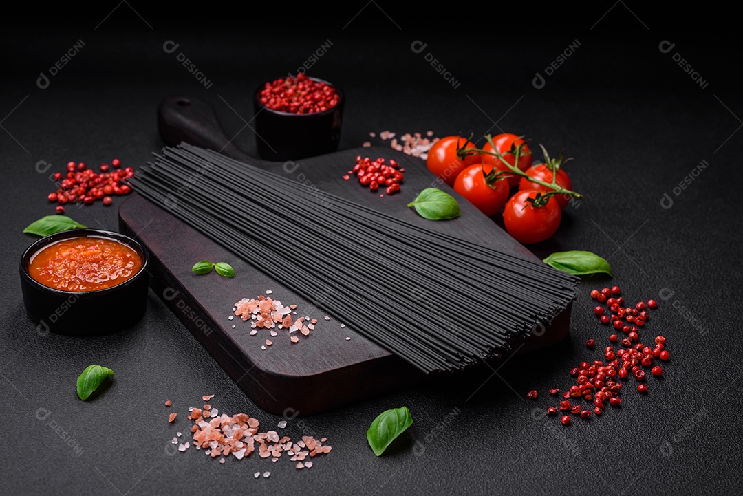 Massa de espaguete preta crua com tomate, sal, especiarias e ervas sobre um fundo escuro de concreto