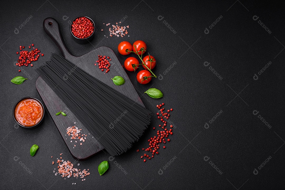 Massa de espaguete preta crua com tomate, sal, especiarias e ervas sobre um fundo escuro de concreto
