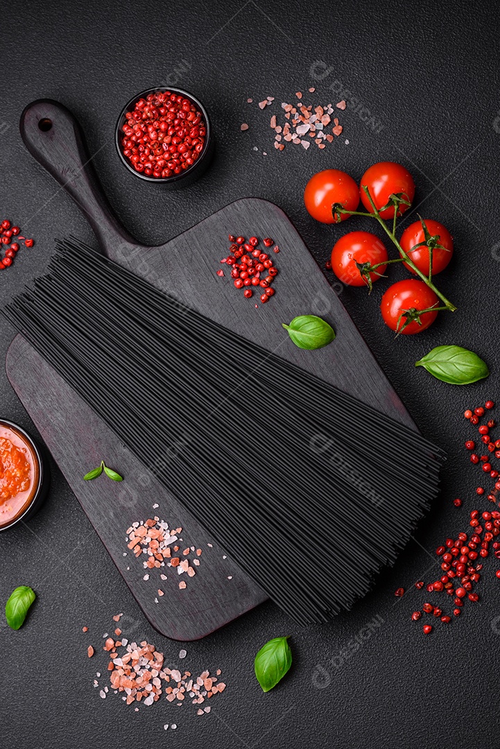 Massa de espaguete preta crua com tomate, sal, especiarias e ervas sobre um fundo escuro de concreto