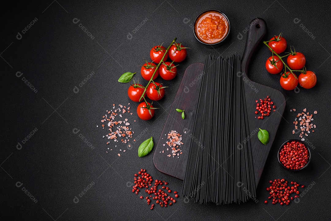 Massa de espaguete preta crua com tomate, sal, especiarias e ervas sobre um fundo escuro de concreto