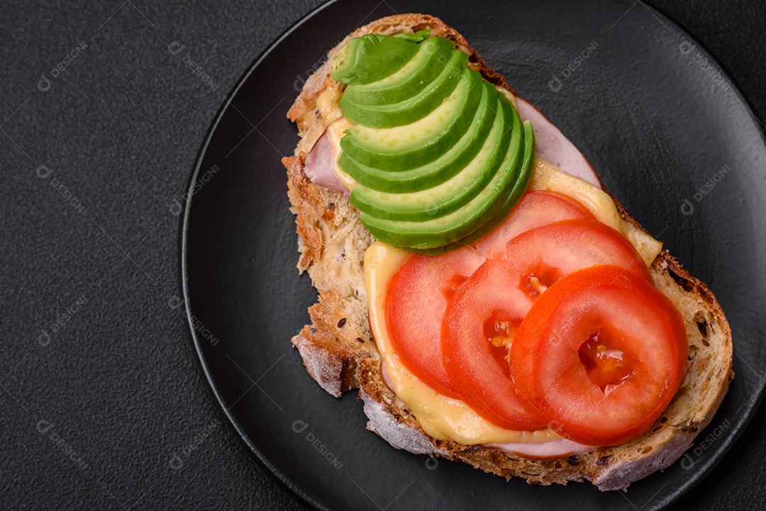 Deliciosa torrada nutritiva grelhada com presunto, queijo, tomate e abacate com sal, especiarias e ervas sobre fundo escuro de concreto