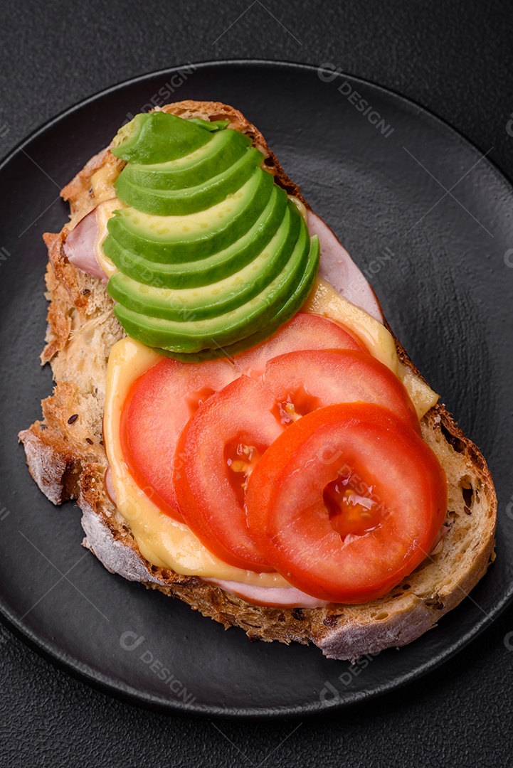 Deliciosa torrada nutritiva grelhada com presunto, queijo, tomate e abacate com sal, especiarias e ervas sobre fundo escuro de concreto