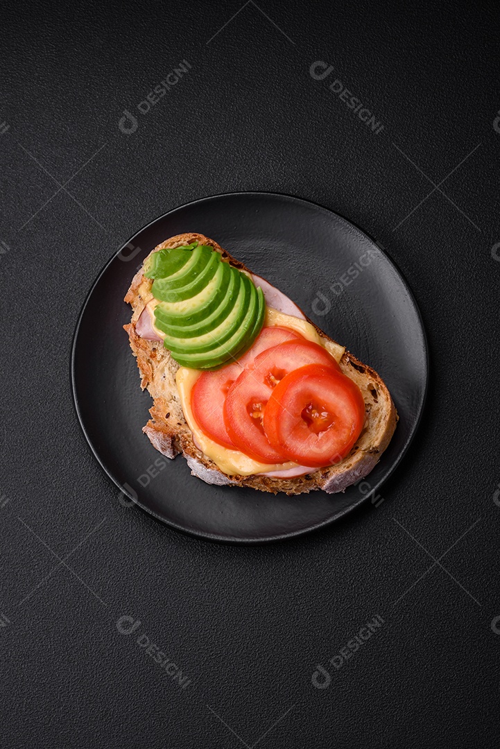 Deliciosa torrada nutritiva grelhada com presunto, queijo, tomate e abacate com sal, especiarias e ervas sobre fundo escuro de concreto