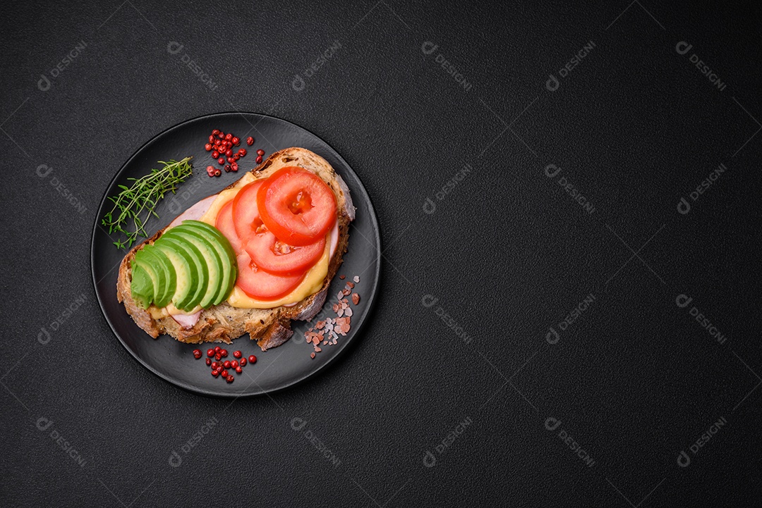 Deliciosa torrada nutritiva grelhada com presunto, queijo, tomate e abacate com sal, especiarias e ervas sobre fundo escuro de concreto