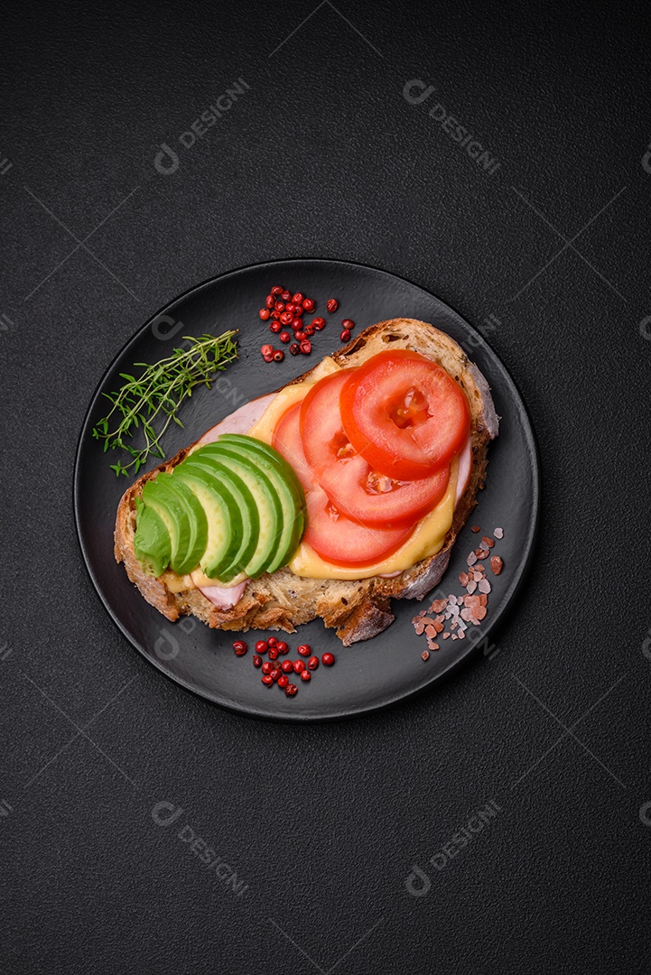 Deliciosa torrada nutritiva grelhada com presunto, queijo, tomate e abacate com sal, especiarias e ervas sobre fundo escuro de concreto