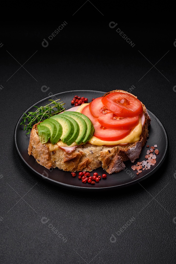 Deliciosa torrada nutritiva grelhada com presunto, queijo, tomate e abacate com sal, especiarias e ervas sobre fundo escuro de concreto