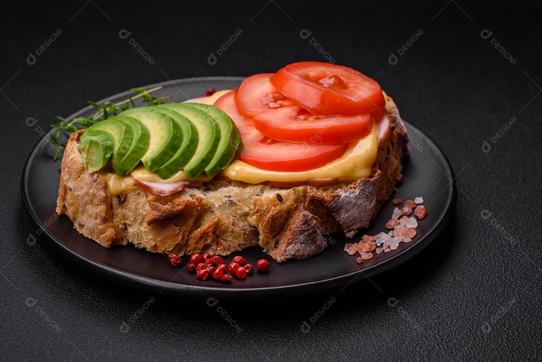 Deliciosa torrada nutritiva grelhada com presunto, queijo, tomate e abacate com sal, especiarias e ervas sobre fundo escuro de concreto