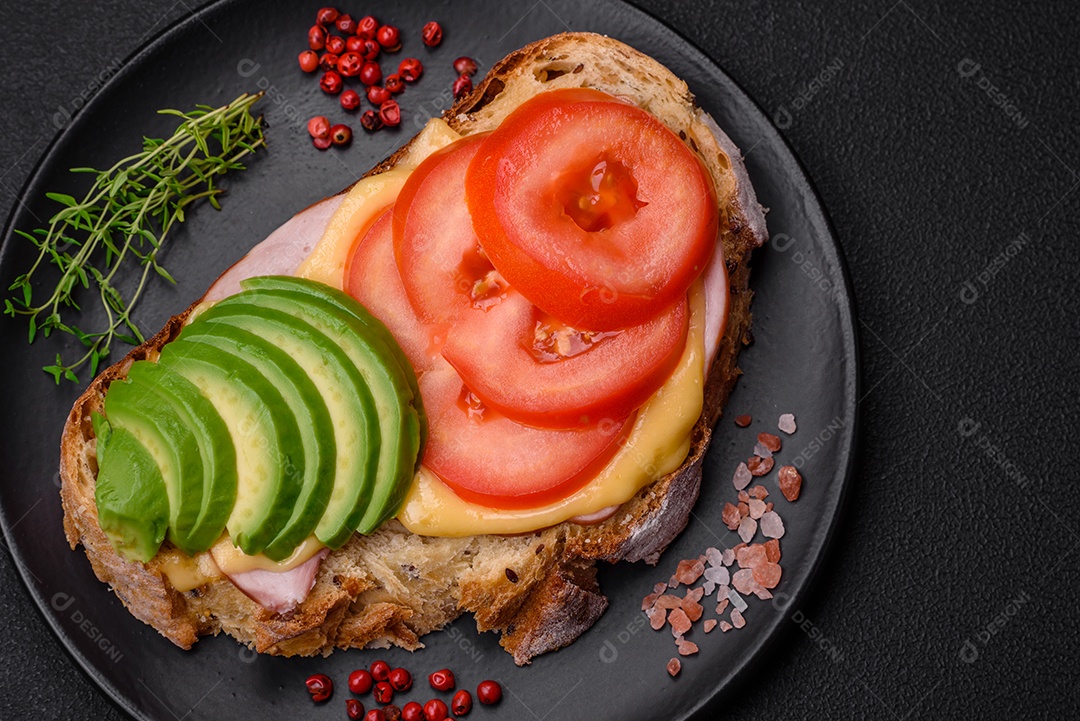 Deliciosa torrada nutritiva grelhada com presunto, queijo, tomate e abacate com sal, especiarias e ervas sobre fundo escuro de concreto