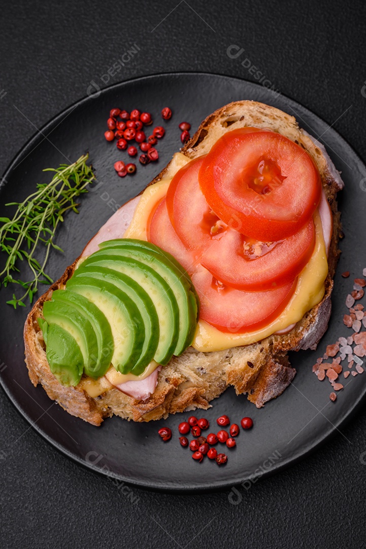 Deliciosa torrada nutritiva grelhada com presunto, queijo, tomate e abacate com sal, especiarias e ervas sobre fundo escuro de concreto