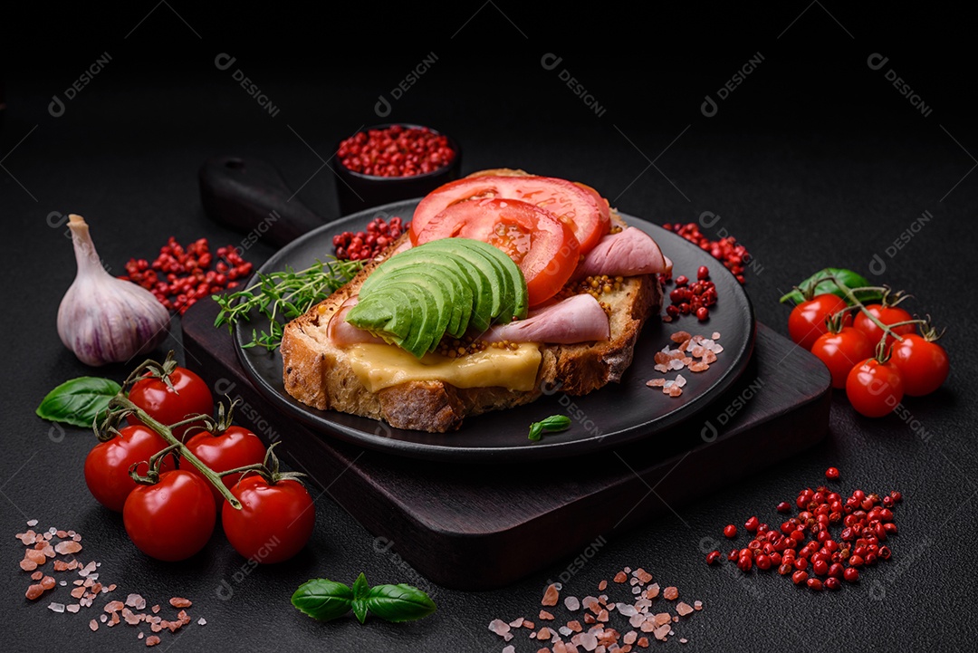 Deliciosa torrada nutritiva grelhada com presunto, queijo, tomate e abacate com sal, especiarias e ervas sobre fundo escuro de concreto