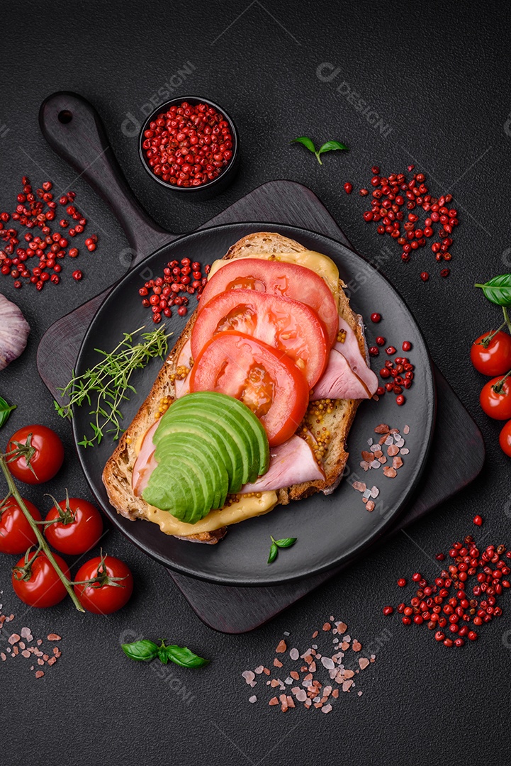 Deliciosa torrada nutritiva grelhada com presunto, queijo, tomate e abacate com sal, especiarias e ervas sobre fundo escuro de concreto