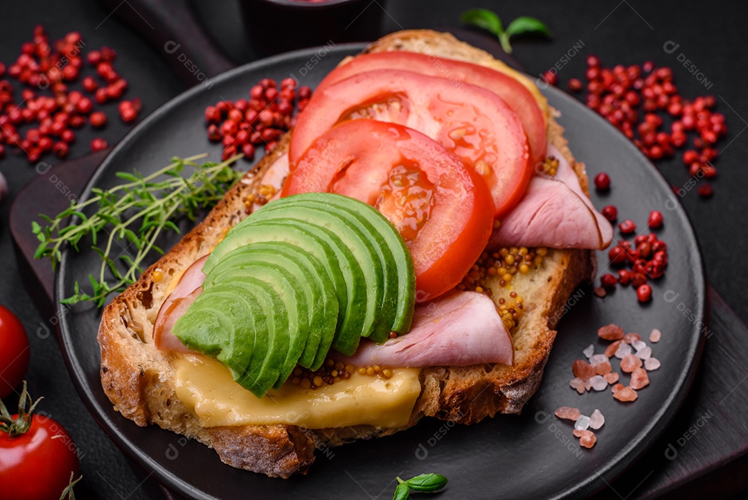 Deliciosa torrada nutritiva grelhada com presunto, queijo, tomate e abacate com sal, especiarias e ervas sobre fundo escuro de concreto