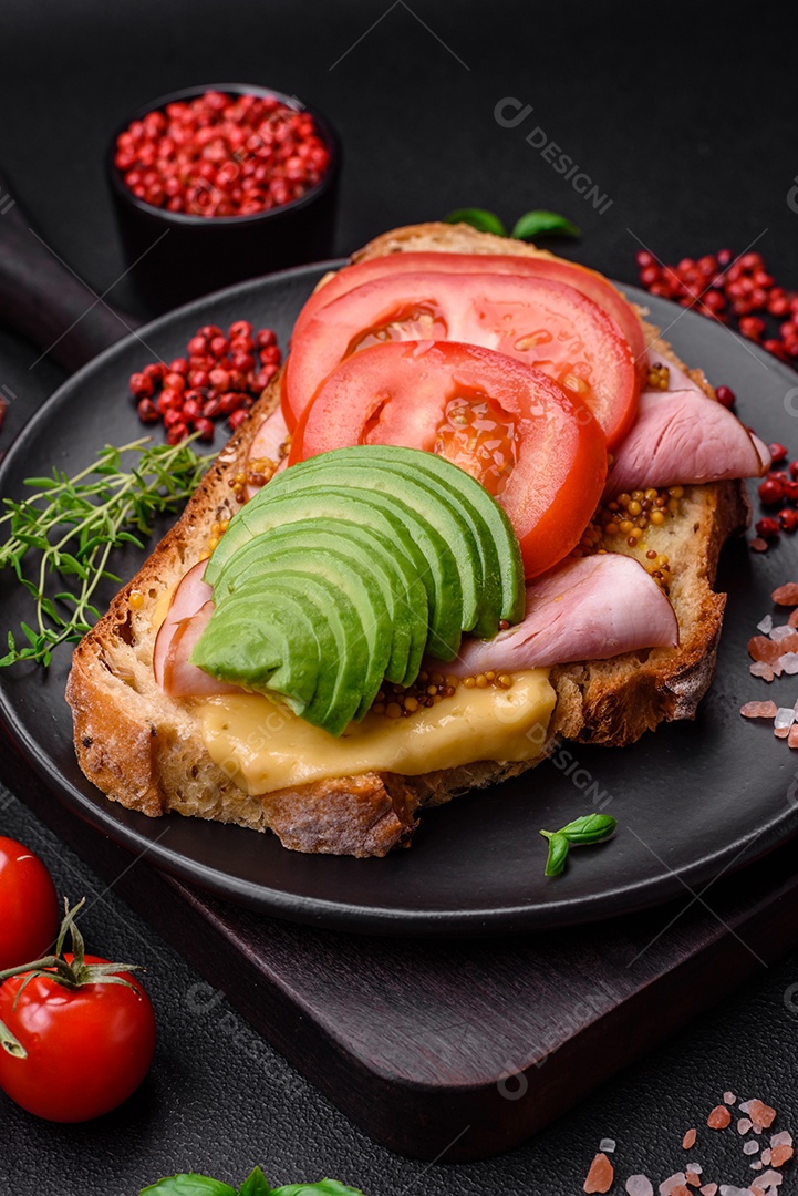 Deliciosa torrada nutritiva grelhada com presunto, queijo, tomate e abacate com sal, especiarias e ervas sobre fundo escuro de concreto