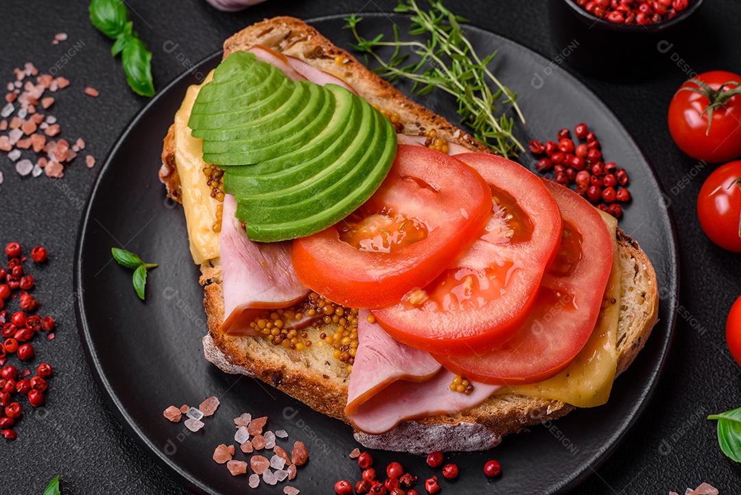 Deliciosa torrada nutritiva grelhada com presunto, queijo, tomate e abacate com sal, especiarias e ervas sobre fundo escuro de concreto