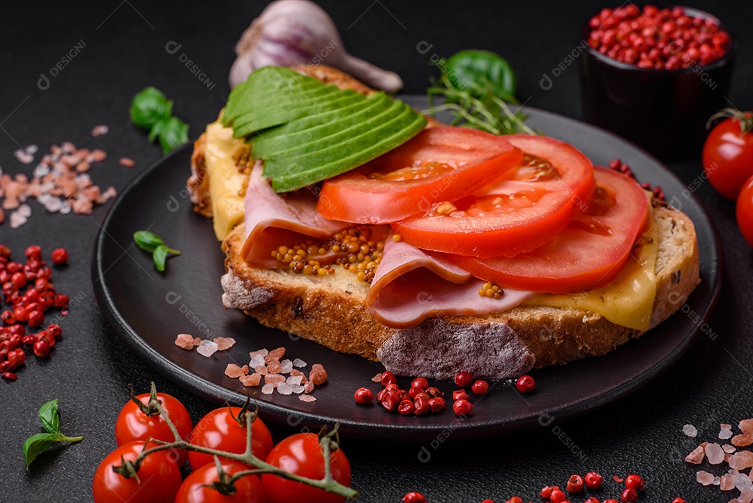 Deliciosa torrada nutritiva grelhada com presunto, queijo, tomate e abacate com sal, especiarias e ervas sobre fundo escuro de concreto