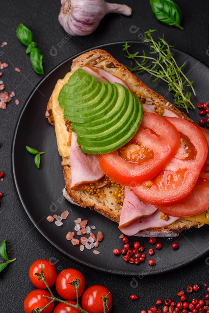 Deliciosa torrada nutritiva grelhada com presunto, queijo, tomate e abacate com sal, especiarias e ervas sobre fundo escuro de concreto