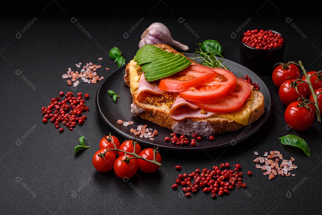 Deliciosa torrada nutritiva grelhada com presunto, queijo, tomate e abacate com sal, especiarias e ervas sobre fundo escuro de concreto