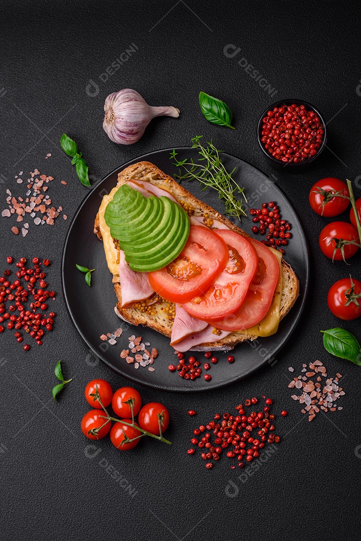 Deliciosa torrada nutritiva grelhada com presunto, queijo, tomate e abacate com sal, especiarias e ervas sobre fundo escuro de concreto