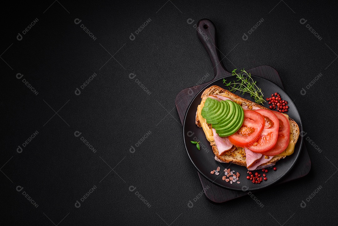 Deliciosa torrada nutritiva grelhada com presunto, queijo, tomate e abacate com sal, especiarias e ervas sobre fundo escuro de concreto