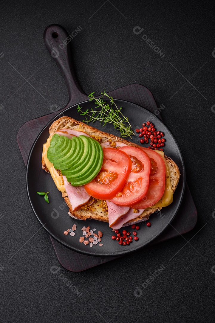 Deliciosa torrada nutritiva grelhada com presunto, queijo, tomate e abacate com sal, especiarias e ervas sobre fundo escuro de concreto
