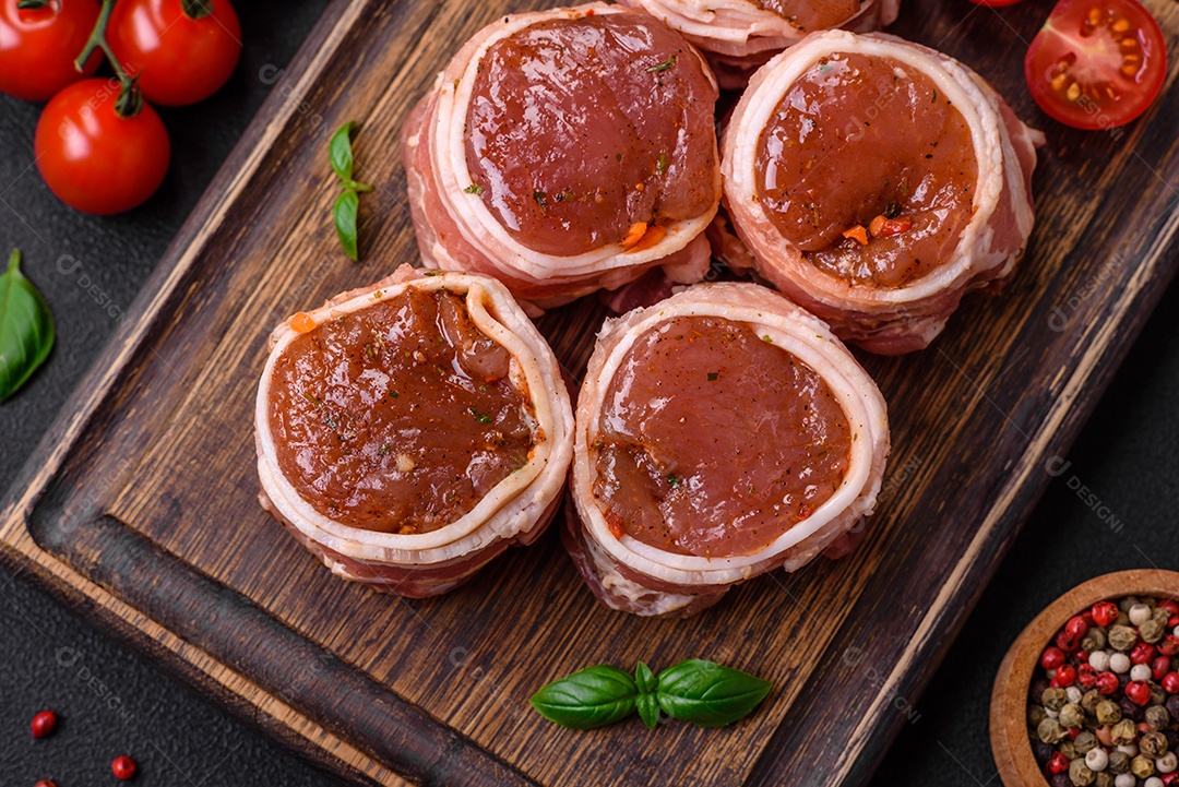 Deliciosos rolos de carne de porco ou frango crus e frescos embrulhados em bacon, com sal, especiarias e ervas sobre um fundo escuro de concreto