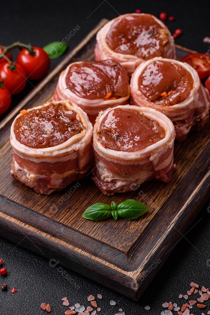 Deliciosos rolos de carne de porco ou frango crus e frescos embrulhados em bacon, com sal, especiarias e ervas sobre um fundo escuro de concreto