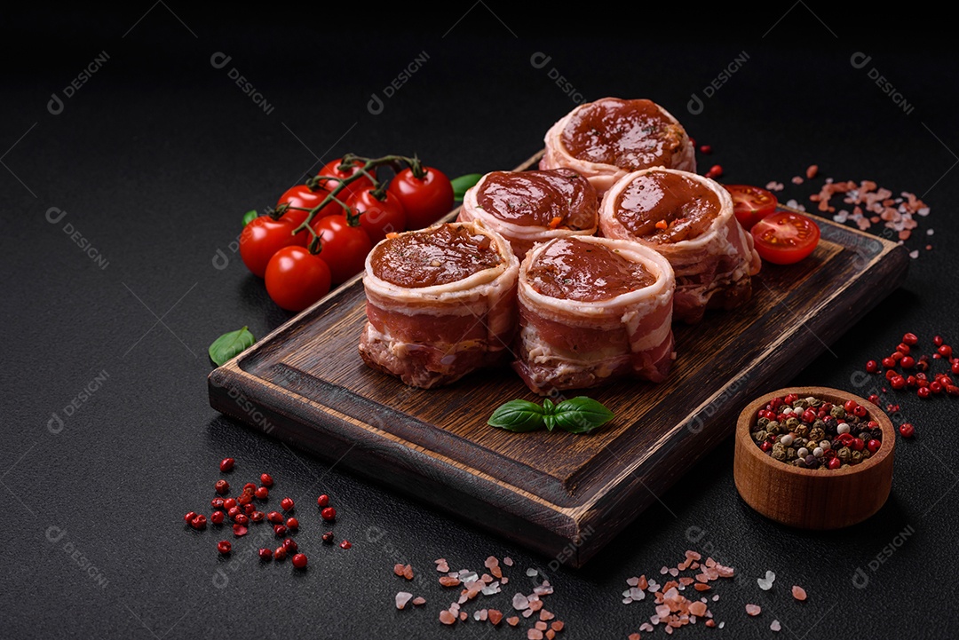 Deliciosos rolos de carne de porco ou frango crus e frescos embrulhados em bacon, com sal, especiarias e ervas sobre um fundo escuro de concreto