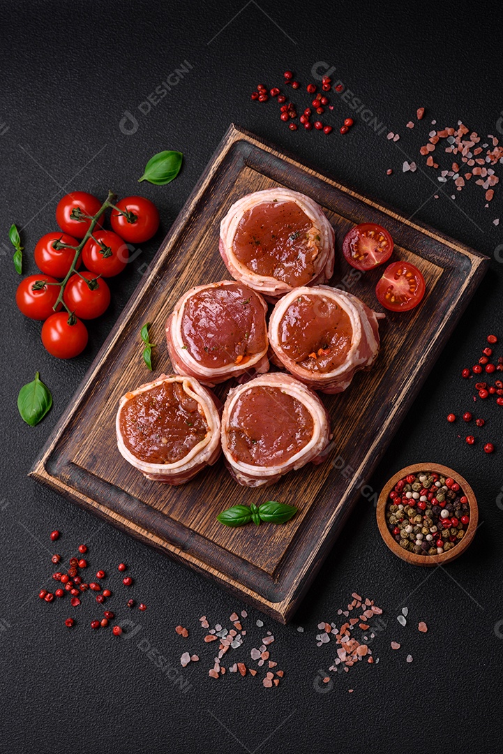 Deliciosos rolos de carne de porco ou frango crus e frescos embrulhados em bacon, com sal, especiarias e ervas sobre um fundo escuro de concreto