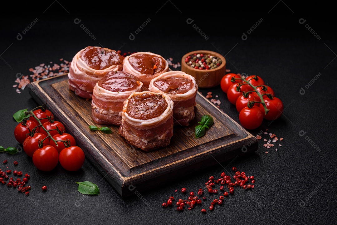 Deliciosos rolos de carne de porco ou frango crus e frescos embrulhados em bacon, com sal, especiarias e ervas sobre um fundo escuro de concreto