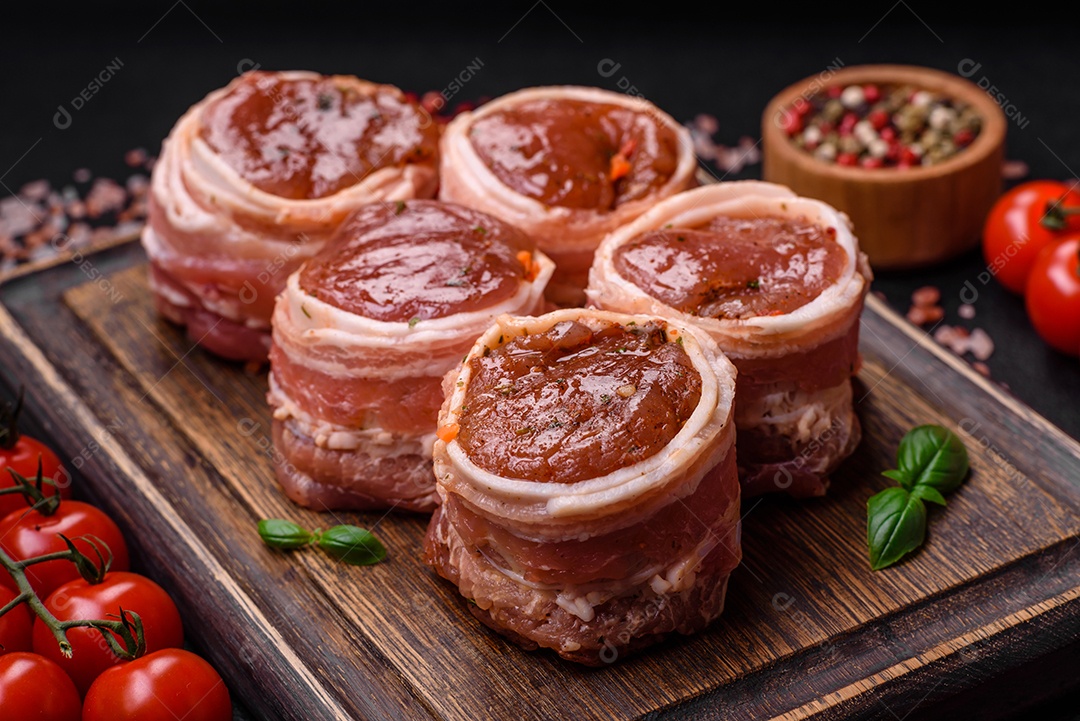 Deliciosos rolos de carne de porco ou frango crus e frescos embrulhados em bacon, com sal, especiarias e ervas sobre um fundo escuro de concreto