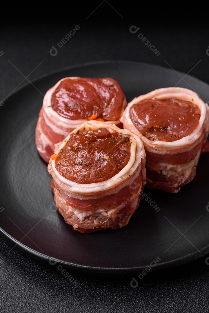 Deliciosos rolos de carne de porco ou frango crus e frescos embrulhados em bacon, com sal, especiarias e ervas sobre um fundo escuro de concreto