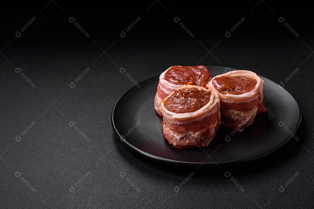 Deliciosos rolos de carne de porco ou frango crus e frescos embrulhados em bacon, com sal, especiarias e ervas sobre um fundo escuro de concreto