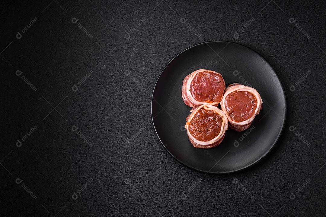 Deliciosos rolos de carne de porco ou frango crus e frescos embrulhados em bacon, com sal, especiarias e ervas sobre um fundo escuro de concreto