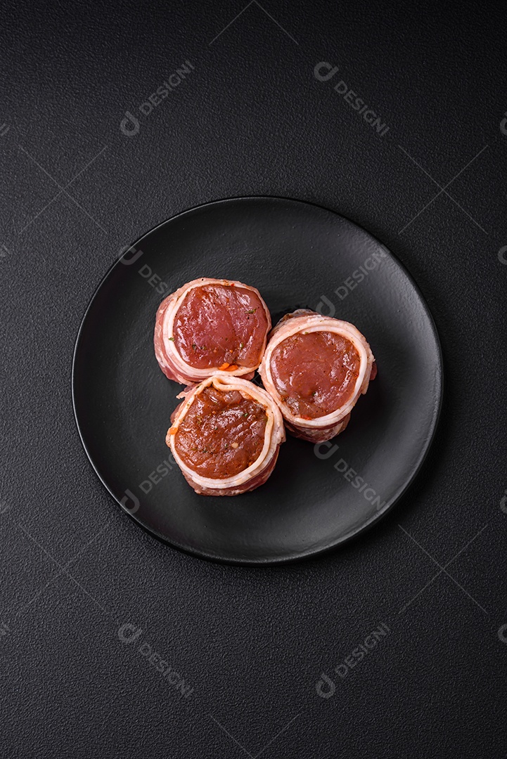 Deliciosos rolos de carne de porco ou frango crus e frescos embrulhados em bacon, com sal, especiarias e ervas sobre um fundo escuro de concreto