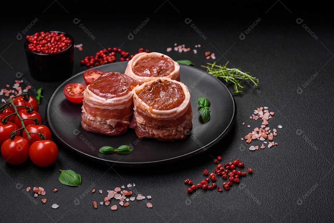 Deliciosos rolos de carne de porco ou frango crus e frescos embrulhados em bacon, com sal, especiarias e ervas sobre um fundo escuro de concreto