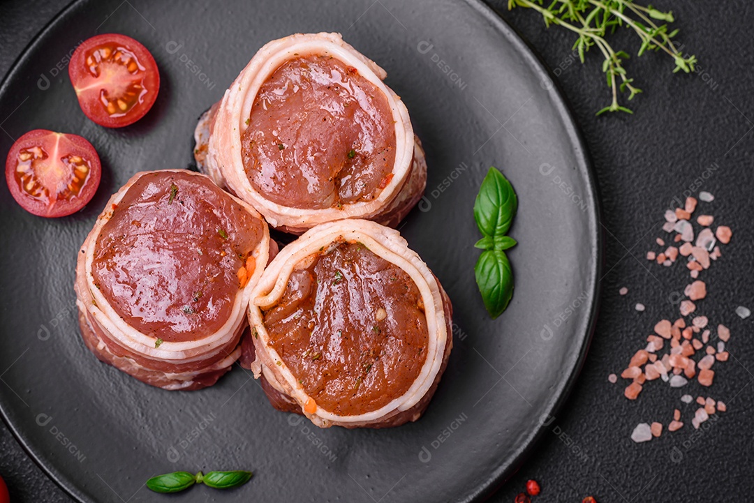 Deliciosos rolos de carne de porco ou frango crus e frescos embrulhados em bacon, com sal, especiarias e ervas sobre um fundo escuro de concreto