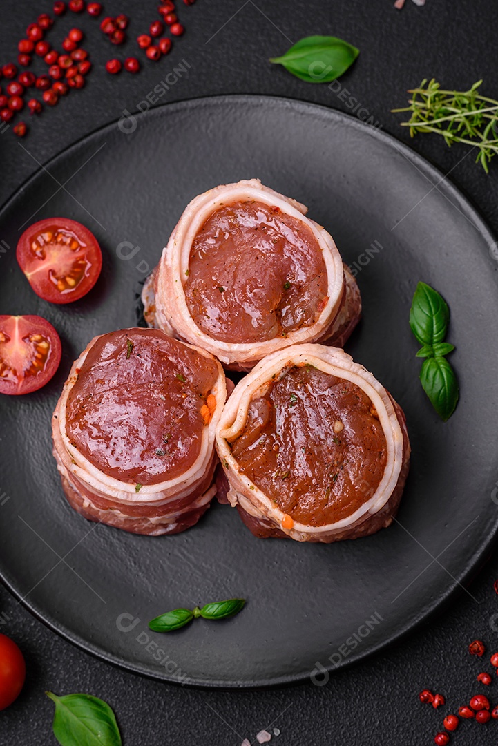 Deliciosos rolos de carne de porco ou frango crus e frescos embrulhados em bacon, com sal, especiarias e ervas sobre um fundo escuro de concreto