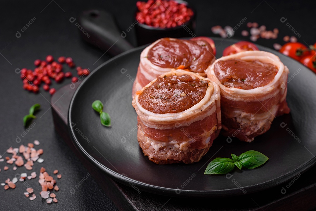 Deliciosos rolos de carne de porco ou frango crus e frescos embrulhados em bacon, com sal, especiarias e ervas sobre um fundo escuro de concreto