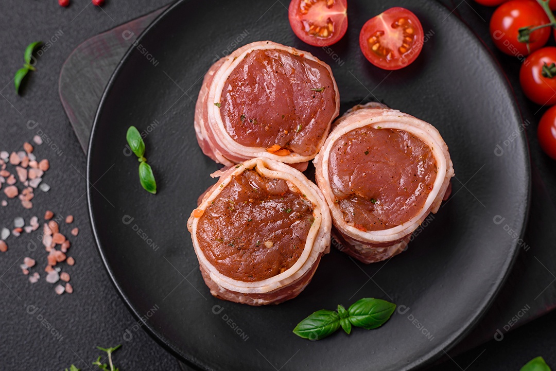 Deliciosos rolos de carne de porco ou frango crus e frescos embrulhados em bacon, com sal, especiarias e ervas sobre um fundo escuro de concreto