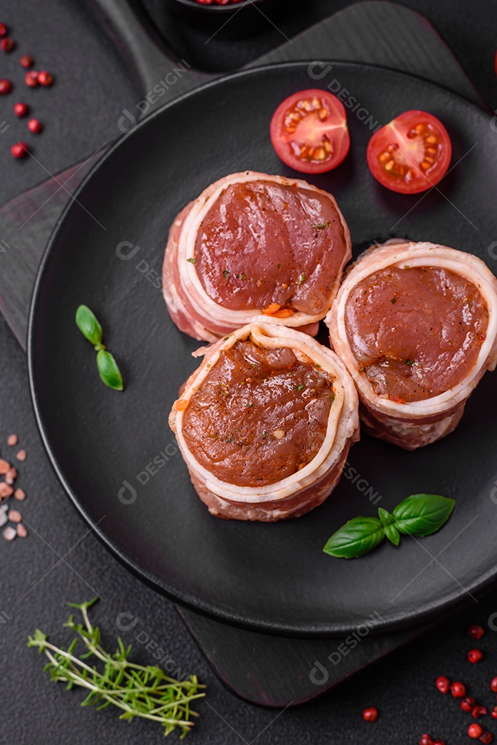 Deliciosos rolos de carne de porco ou frango crus e frescos embrulhados em bacon, com sal, especiarias e ervas sobre um fundo escuro de concreto