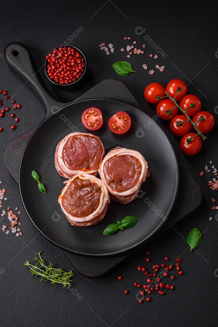 Deliciosos rolos de carne de porco ou frango crus e frescos embrulhados em bacon, com sal, especiarias e ervas sobre um fundo escuro de concreto