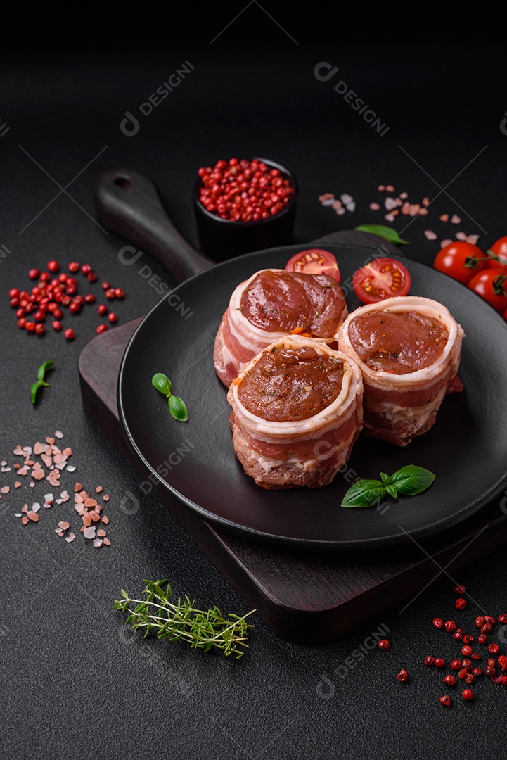 Deliciosos rolos de carne de porco ou frango crus e frescos embrulhados em bacon, com sal, especiarias e ervas sobre um fundo escuro de concreto