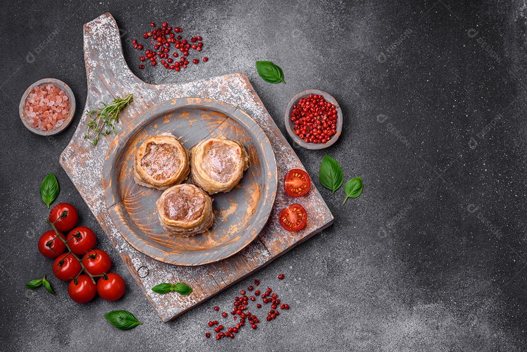 Deliciosos rolos de carne de porco ou frango crus e frescos embrulhados em bacon, com sal, especiarias e ervas sobre um fundo escuro de concreto